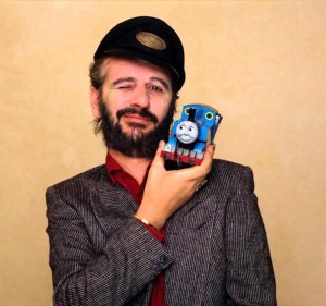 Image of Ringo Starr wearing a conductor hat, winking, and holding up a Thomas the Tank Engine toy against a beige background